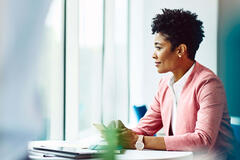 Womale sitting at a table with phone in hand. Tablet and notebook on table. Womale looking out of the window.