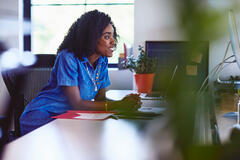 Une femme souriante en robe bleue est assise à son bureau. Elle regarde un écran. Une plante verte et un dossier rouge apportent de la couleur à cet espace de travail lumineux.