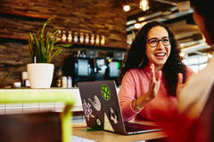 Une femme à lunettes rit en discutant devant son ordinateur portable dans un café chaleureux. Elle porte un pull rose. Une plante et un bar en bois se trouvent en arrière-plan.