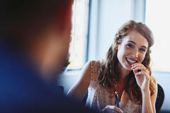 Une femme souriante discute avec un collègue au bureau. Elle tient un stylo, prête à noter. L'ambiance est lumineuse et professionnelle, illustrant une collaboration sereine.