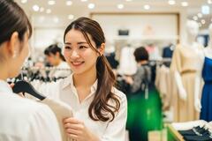 Une femme souriante en chemisier blanc conseille une cliente dans un magasin de vêtements lumineux. Des mannequins et d'autres articles de mode sont visibles en arrière-plan.