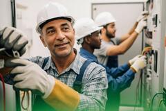  3 techniciens en casque blanc et gants de protection travaillent sur une armoire électrique dans une station nucléaire. L'homme au premier plan sourit, illustrant un environnement de travail collaboratif et sécurisé.