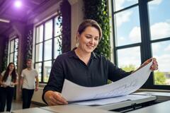 Une femme souriante en chemise noire examine un grand plan technique. Elle se trouve dans un bureau lumineux aux grandes fenêtres décorées de plantes. Deux collègues marchent en arrière-plan.