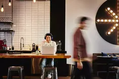Man with headphones working on laptop at a table in a cafe while a man walks by