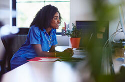 Une femme souriante en robe bleue est assise à son bureau. Elle regarde un écran. Une plante verte et un dossier rouge apportent de la couleur à cet espace de travail lumineux.