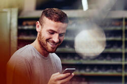 Un homme barbu sourit en consultant son smartphone. Il se trouve dans un atelier ou un entrepôt avec des étagères industrielles en arrière-plan. L'ambiance est positive et détendue.