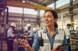 Une femme souriante avec une radio et un gilet de sécurité dirige une équipe dans un hangar aéronautique. Des techniciens s'activent en arrière-plan sous une structure industrielle vaste.