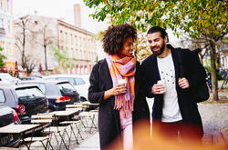 Un homme et une femme marchent en souriant dans une rue, un café à la main. Ils portent des manteaux et une écharpe colorée. 