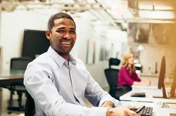 Un homme en chemise bleue sourit à son bureau, les mains sur son clavier. En arrière-plan, une collègue en haut rose travaille à son poste dans un open space lumineux.