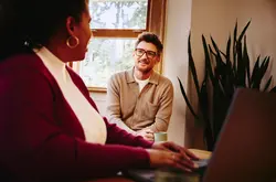 Un homme à lunettes et une femme échangent dans un bureau lumineux, devant une fenêtre et une plante. Elle est au premier plan, dos à nous, travaillant sur un ordinateur portable.