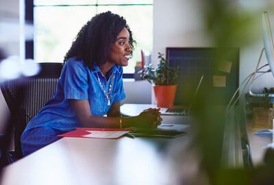 Une femme souriante en robe bleue est assise à son bureau. Elle regarde un écran. Une plante verte et un dossier rouge apportent de la couleur à cet espace de travail lumineux.