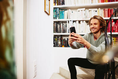 Un homme souriant en pull vert est assis dans un escalier. Il tient son téléphone devant lui. Derrière lui, une grande bibliothèque remplie de livres décore le mur blanc.