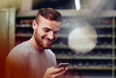 Un homme barbu sourit en consultant son smartphone. Il se trouve dans un atelier ou un entrepôt avec des étagères industrielles en arrière-plan. L'ambiance est positive et détendue.