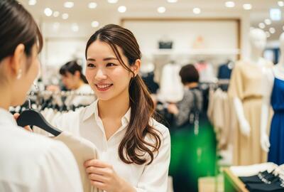 Une femme souriante en chemisier blanc conseille une cliente dans un magasin de vêtements lumineux. Des mannequins et d'autres articles de mode sont visibles en arrière-plan.