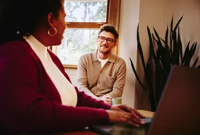 Un homme à lunettes et une femme échangent dans un bureau lumineux, devant une fenêtre et une plante. Elle est au premier plan, dos à nous, travaillant sur un ordinateur portable.