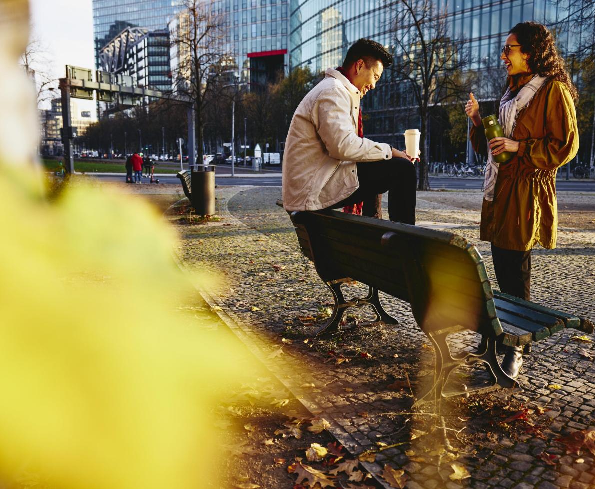 Man and woman holding drinks smiling and having a conversation. Man is sitting down on bench.