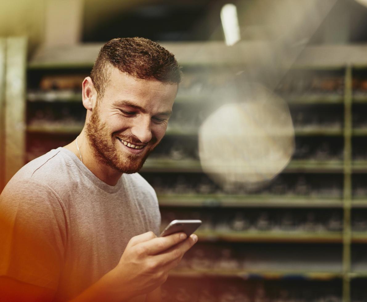 Blue-collar worker looking at his mobile phone. Tech environment. Smiling. Groomed beard. Greyish T-shirt. Primary color red. travailleur regardant son téléphone