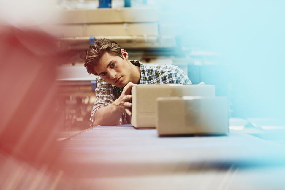 Male blue-collar worker checking parcels. Caucasian man. Checkered shirt. Smooth-shaven chin. Conveyor belt. Logistics environment. Primary color light-red. Secondary colors light- blue and cream.